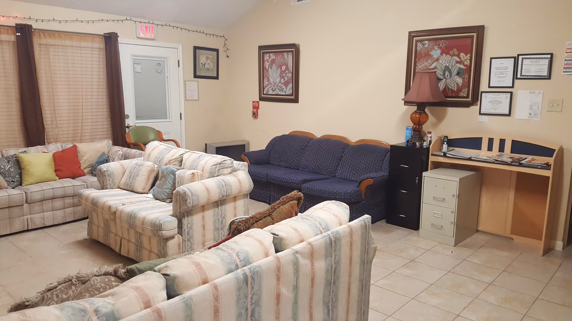 Communal living room with multiple upholstered sofas and chairs, a small desk with a lamp and framed artwork on the walls.