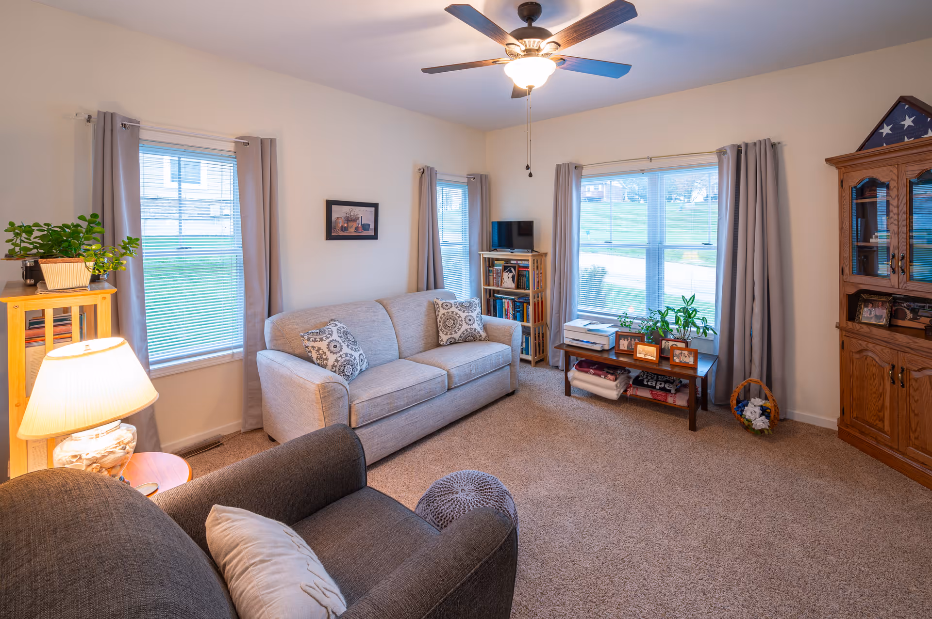 A cozy living room with a beige sofa, a dark gray armchair, a wooden bookshelf, a wooden cabinet with glass doors, and a coffee table with plants and framed photos. The room has beige carpet, three windows with gray curtains, a ceiling fan with light, and a table lamp on a small wooden stand.