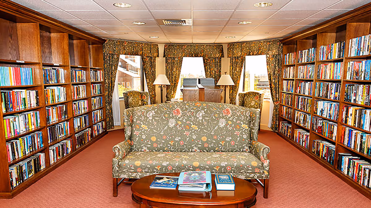A cozy library room with wooden bookshelves filled with books lining both walls. In the center, there is a floral-patterned sofa facing a wooden coffee table with several books on it. The room has three windows with floral curtains, two armchairs, and two floor lamps near the windows providing warm lighting.