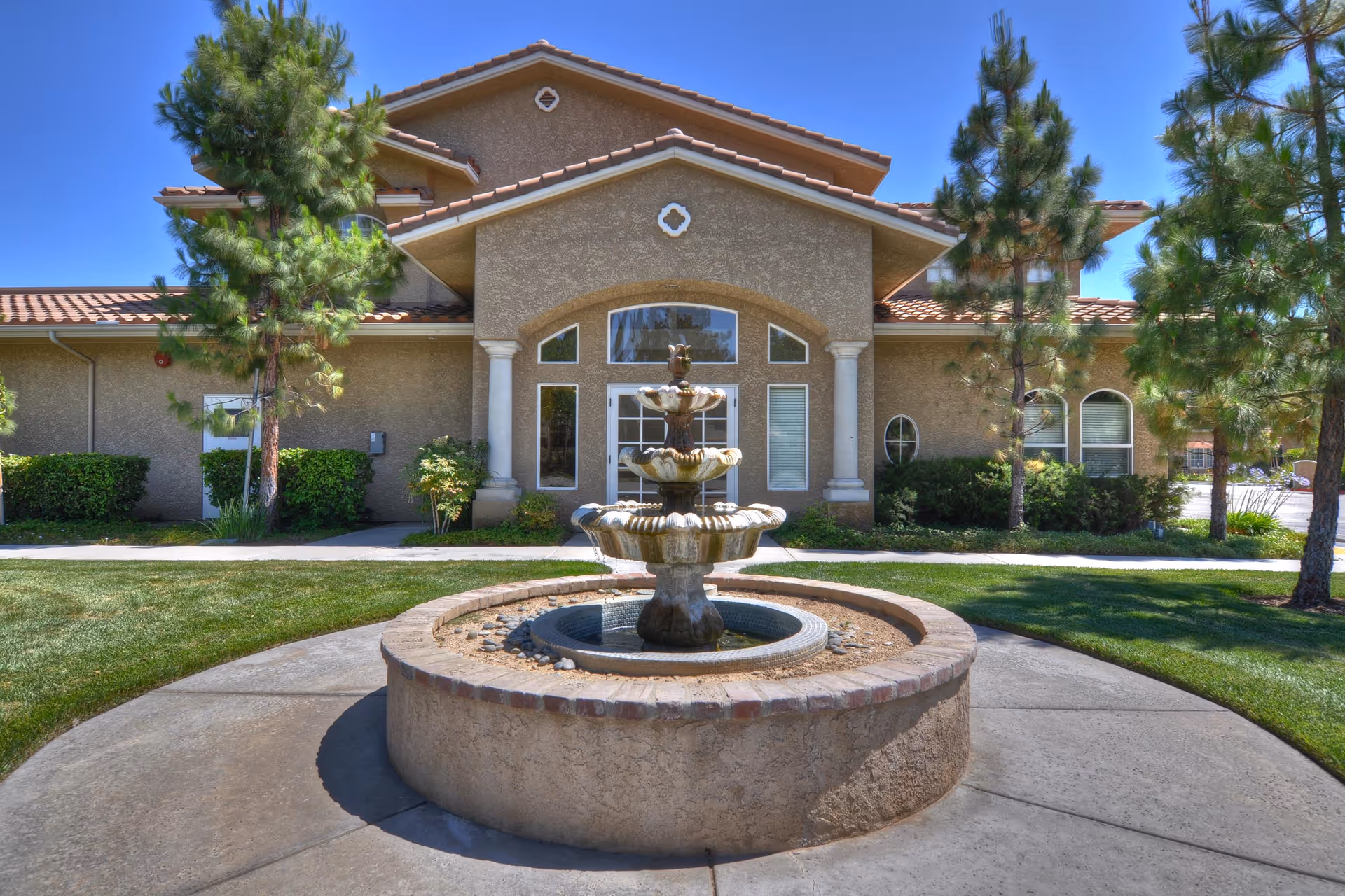 Front exterior view of a building with a beige stucco finish and a tiled roof, featuring a three-tiered stone fountain in the foreground surrounded by a circular stone base. The building has large windows and columns flanking the entrance, with green trees and shrubs on either side under a clear blue sky.