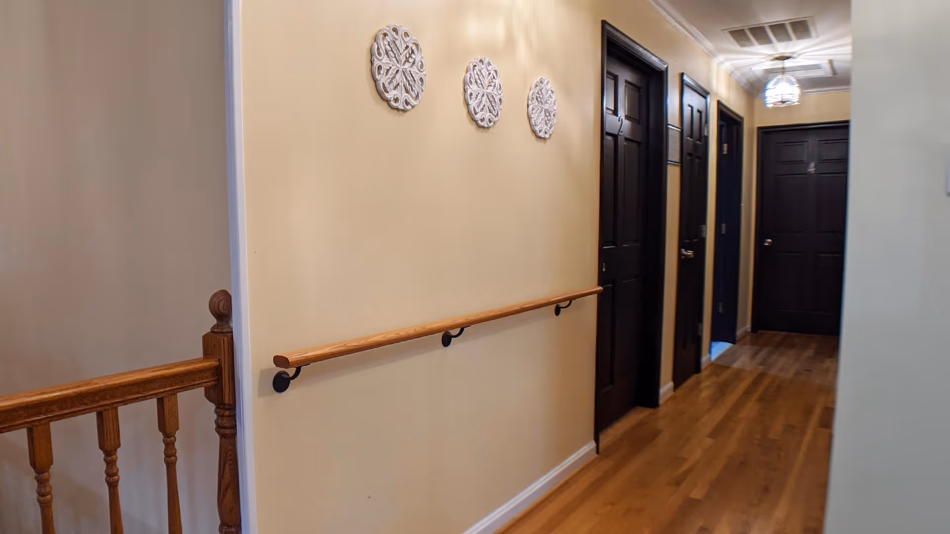 Interior hallway with wooden floor and dark brown doors on the right side. A wooden handrail is mounted on the left wall, which is decorated with three white ornamental wall hangings. A wooden railing is visible on the left side near the foreground. The hallway is lit by a ceiling light fixture.