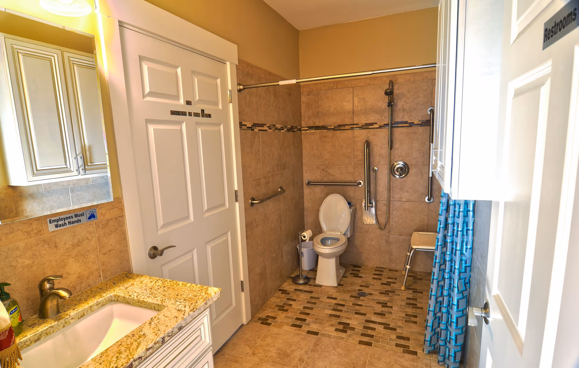 A clean and accessible bathroom with beige tiled walls and floor. The bathroom features a toilet with grab bars around it, a shower area with a handheld showerhead and grab bars, a small shower chair, a granite countertop with a sink, and a mirror above the sink. There are signs on the doors and wall, including one that says 'Employees Must Wash Hands' and another indicating 'Restrooms'.