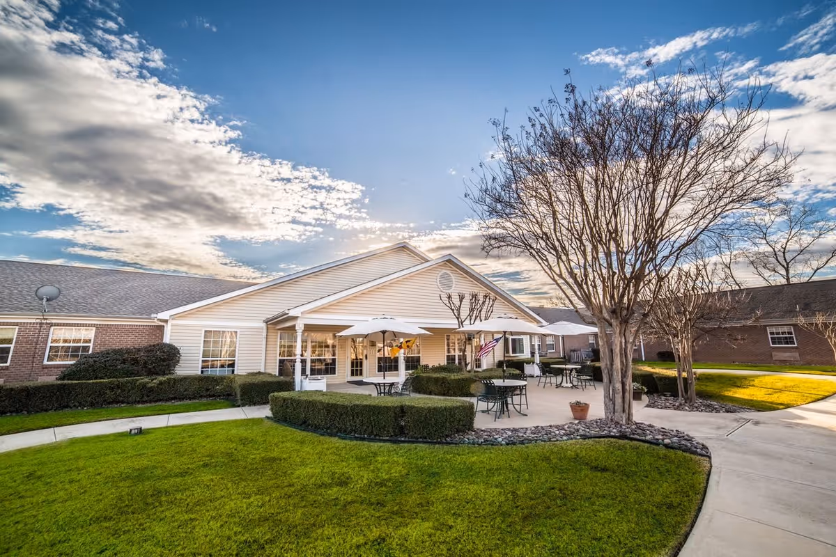 Exterior view of a memory care building with a patio area featuring tables, umbrellas, trimmed hedges, trees and a green lawn under a partly cloudy sky.