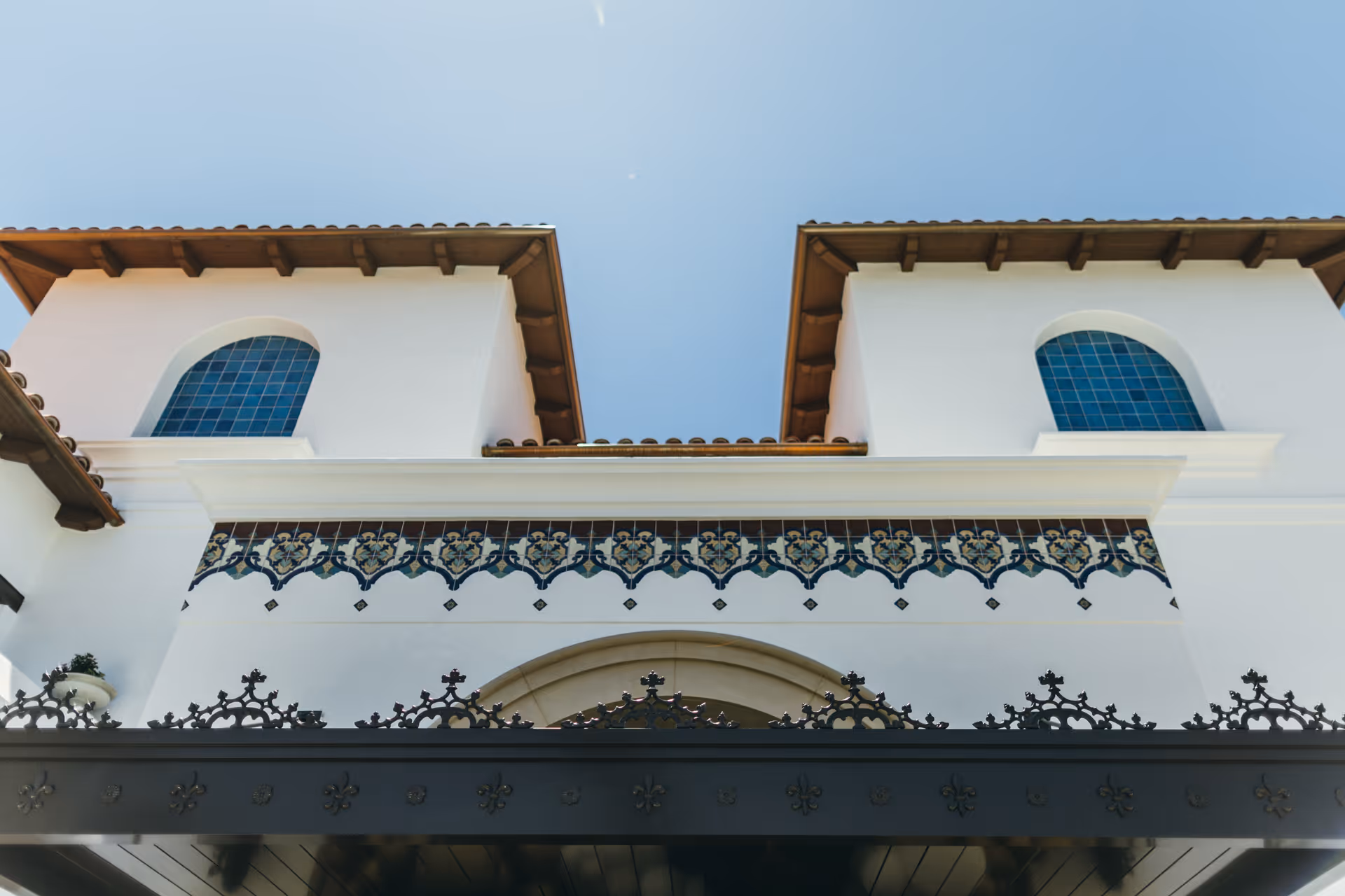 Front exterior of a white Mediterranean-style building with two arched blue-tiled windows, a decorative tile band and a wrought-iron canopy against a clear sky.