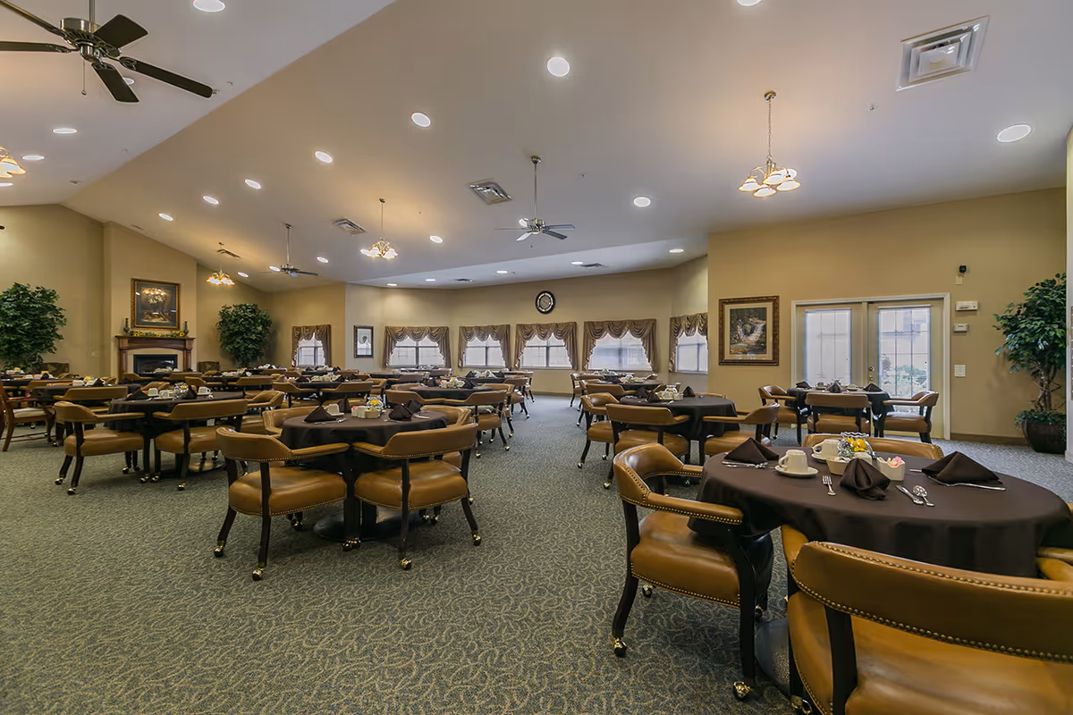 Spacious dining room with round tables covered in dark tablecloths, brown upholstered chairs, chandeliers, and windows along the far wall.