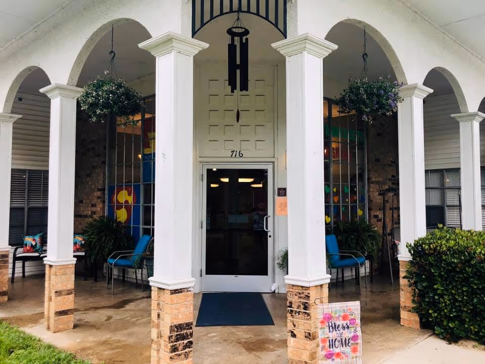 Front entrance with white columns and an arched portico leading to a glass door, flanked by chairs, hanging plants, and a decorative sign.
