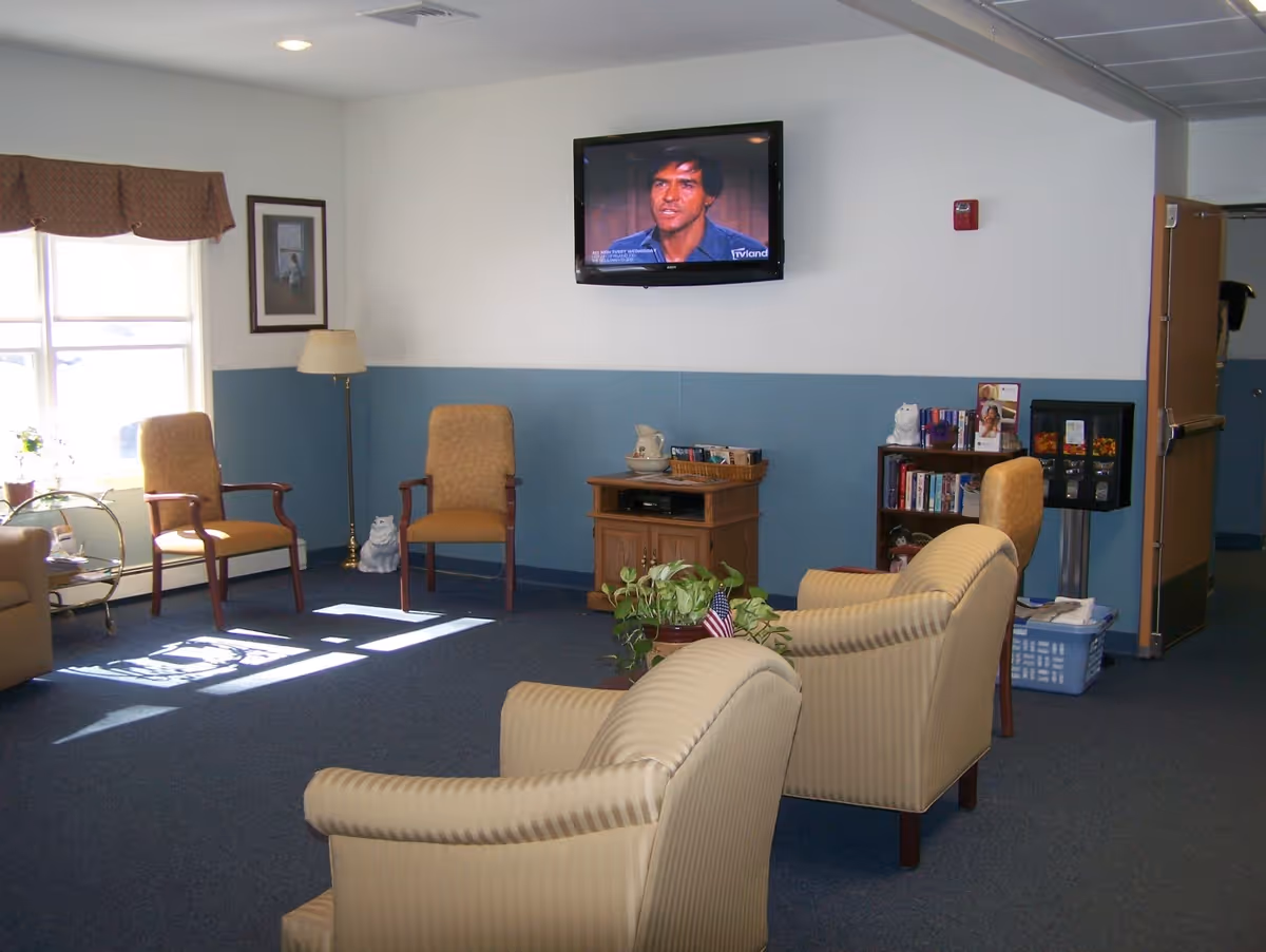 A cozy living room area with beige armchairs and wooden chairs arranged around a small wooden cabinet. A flat-screen TV is mounted on the wall showing a man speaking. The room has blue carpet, white and blue walls, a window with a brown valance, a floor lamp, a small bookshelf with books and decorations, and a potted plant on the floor.