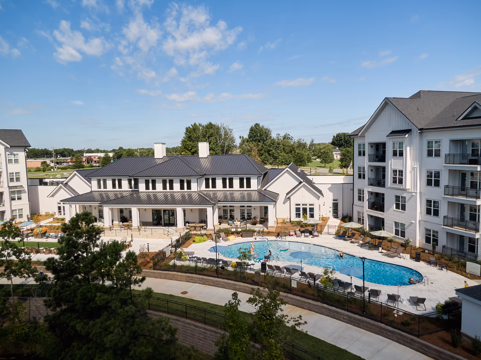 Aerial view of Inspire Royal Park senior living facility showing a large outdoor swimming pool surrounded by lounge chairs and umbrellas, adjacent to a white multi-story building and a clubhouse under a blue sky with scattered clouds.