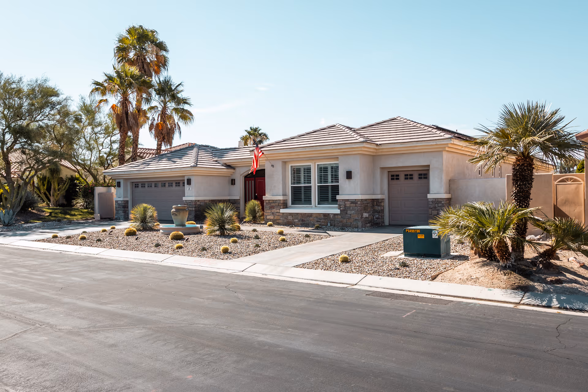 Single-story residential building with a tiled roof, two garages, and a front yard landscaped with desert plants including palm trees and small cacti. The driveway and sidewalk are visible, and an American flag is mounted near the front entrance.