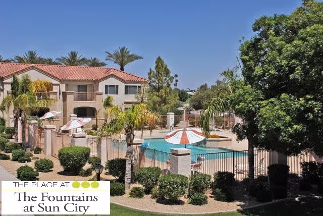 Outdoor view of a senior living facility with a swimming pool surrounded by a fence, palm trees, and shrubs. Two-story beige buildings with red tile roofs are visible in the background under a clear blue sky.