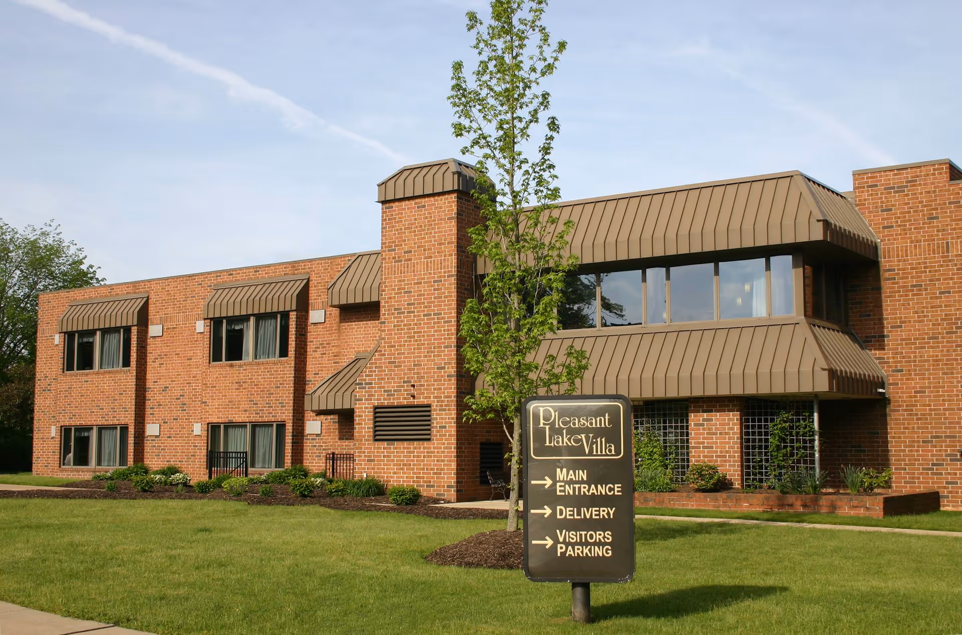 Exterior view of a two-story brick building with brown awnings over the windows and a small tree in front. A sign in the foreground reads Pleasant Lake Villa with directions to the main entrance, delivery, and visitors parking. The building is surrounded by green grass and some shrubs.