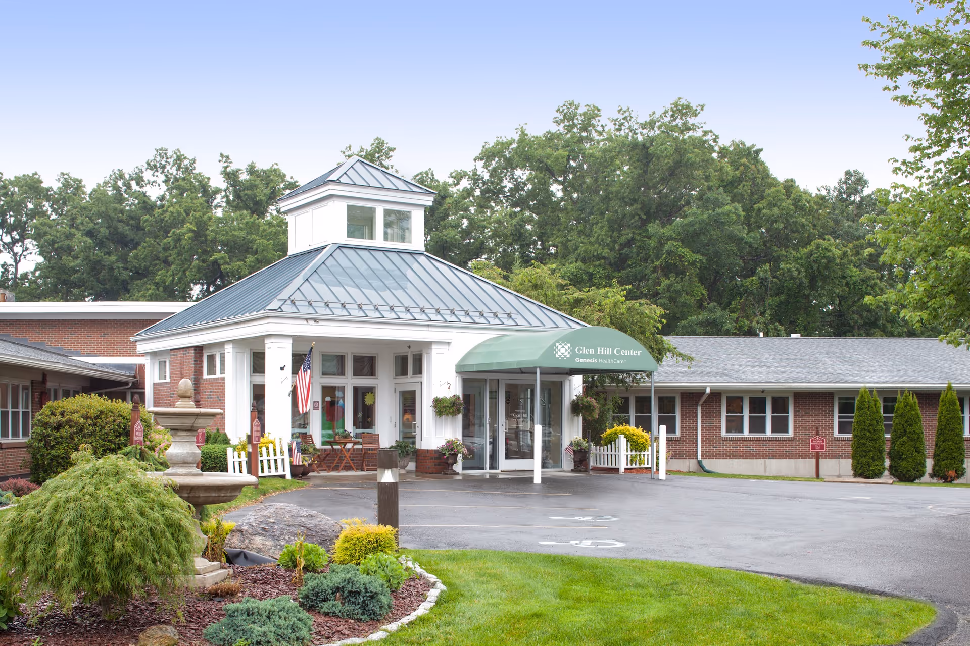 Exterior view of Glen Hill Center, a single-story brick building with a metal roof and a green awning over the entrance. The entrance area has white columns and an American flag displayed. There is a landscaped garden with shrubs and a stone fountain in the foreground, and trees in the background.