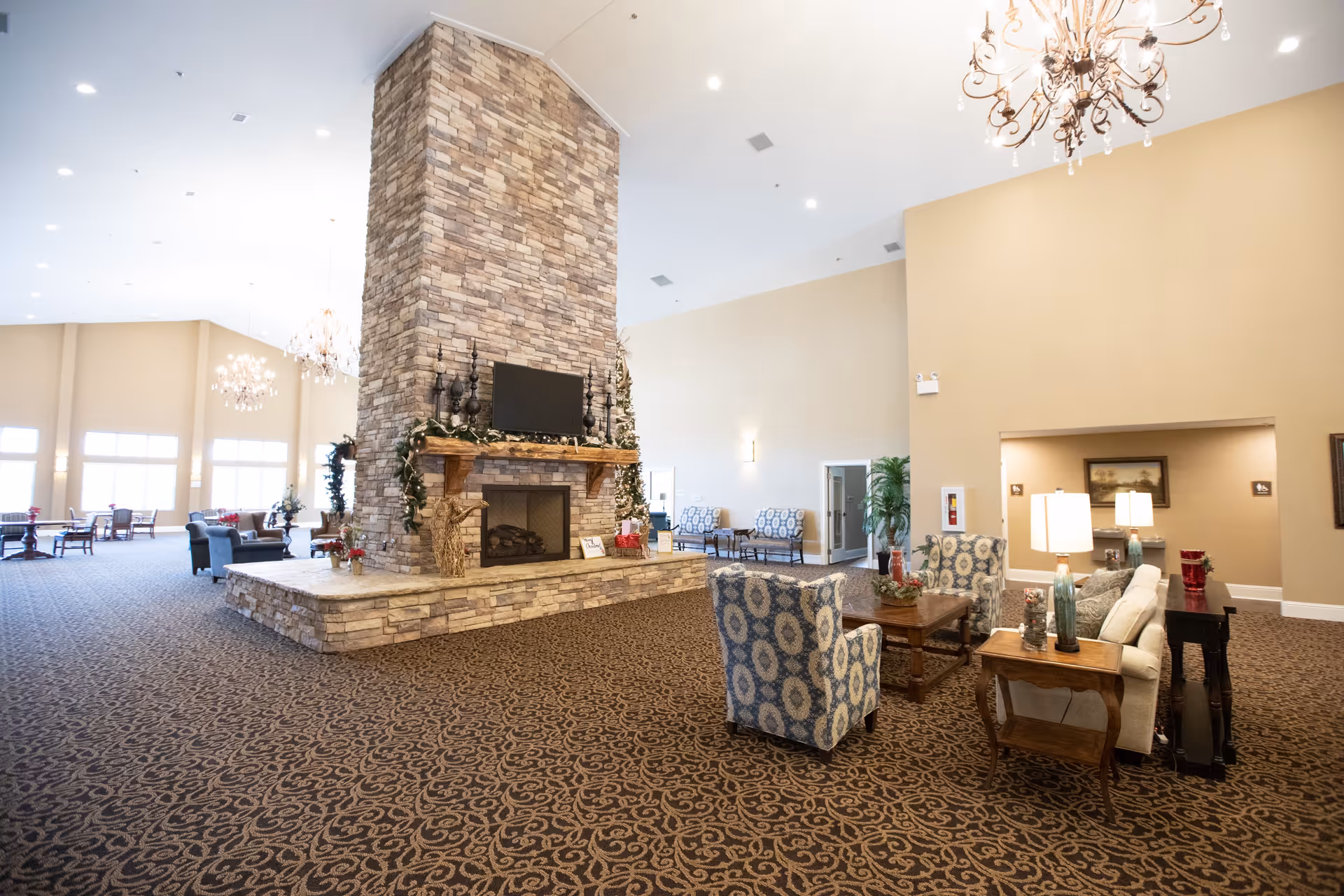 Spacious senior living facility common area with a large stone fireplace in the center, decorated with garlands and a Christmas tree. Comfortable seating including patterned armchairs and sofas arranged around wooden tables with lamps. The room features high ceilings with chandeliers and large windows letting in natural light.