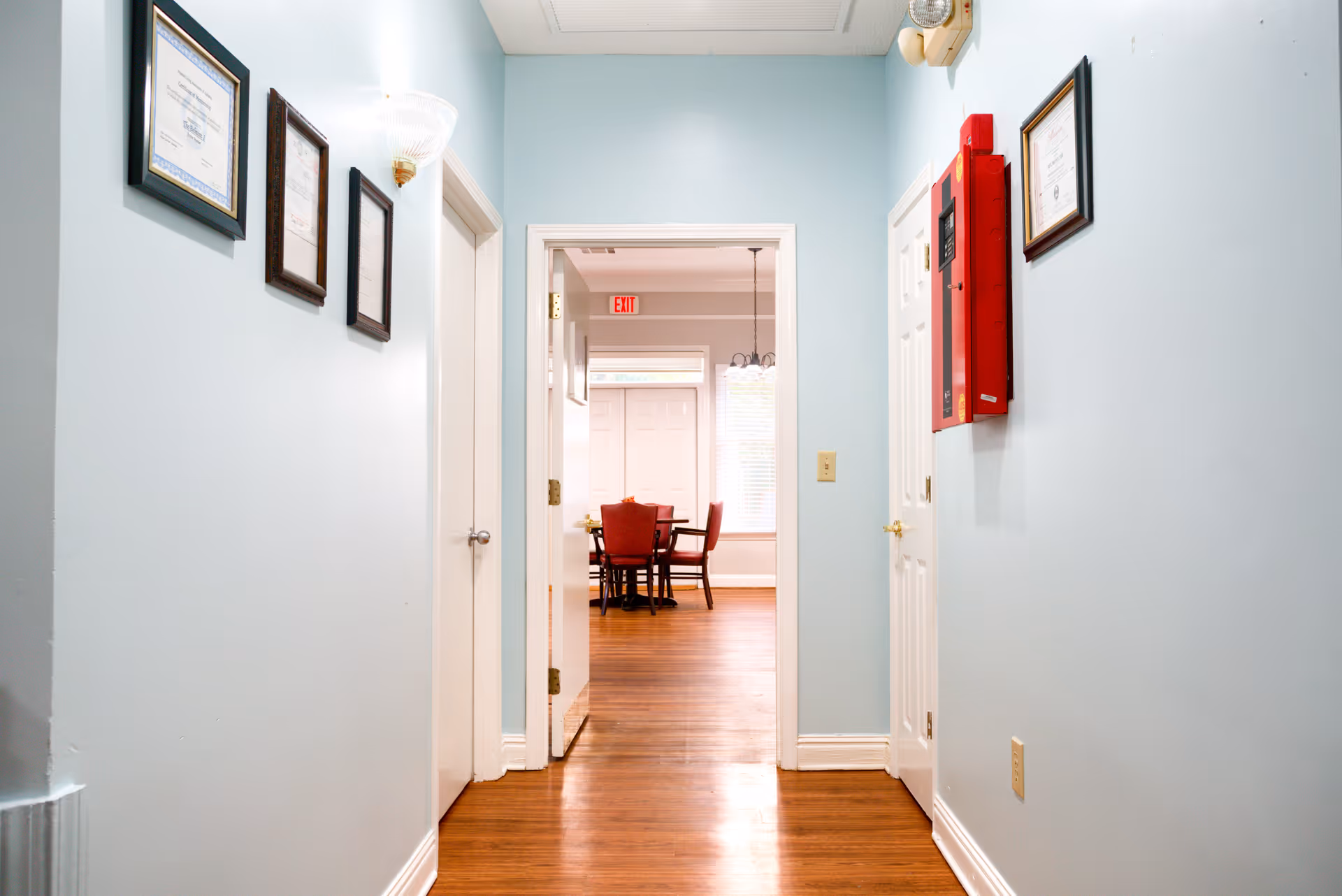 A hallway with light blue walls and wooden floor leading to a room with a table and red chairs. The hallway has framed certificates on the walls and a red fire alarm box on the right side.