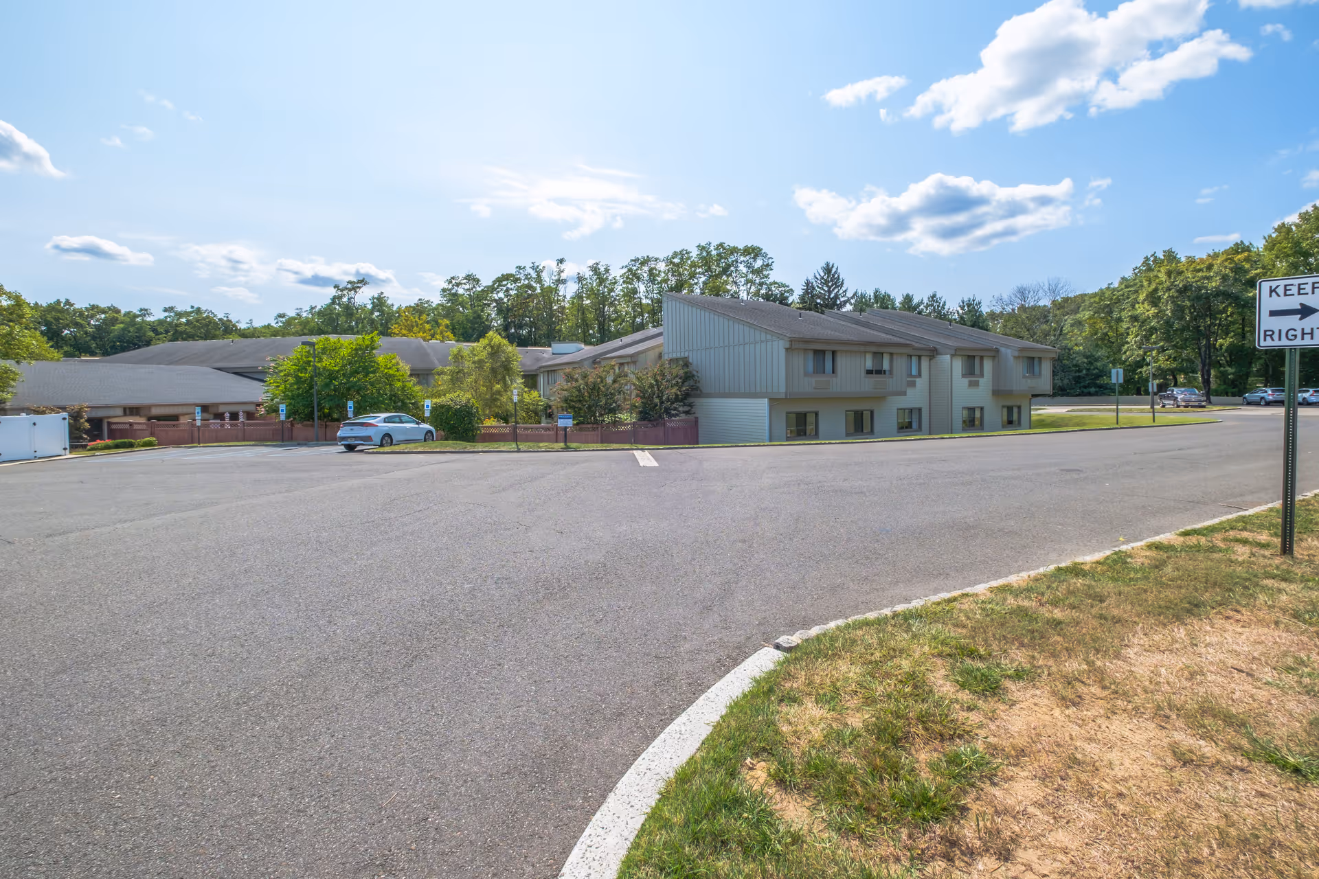 Exterior view of a senior living facility building with a large paved parking area in front. The building has a light-colored facade with multiple windows and is surrounded by trees and greenery under a partly cloudy sky.