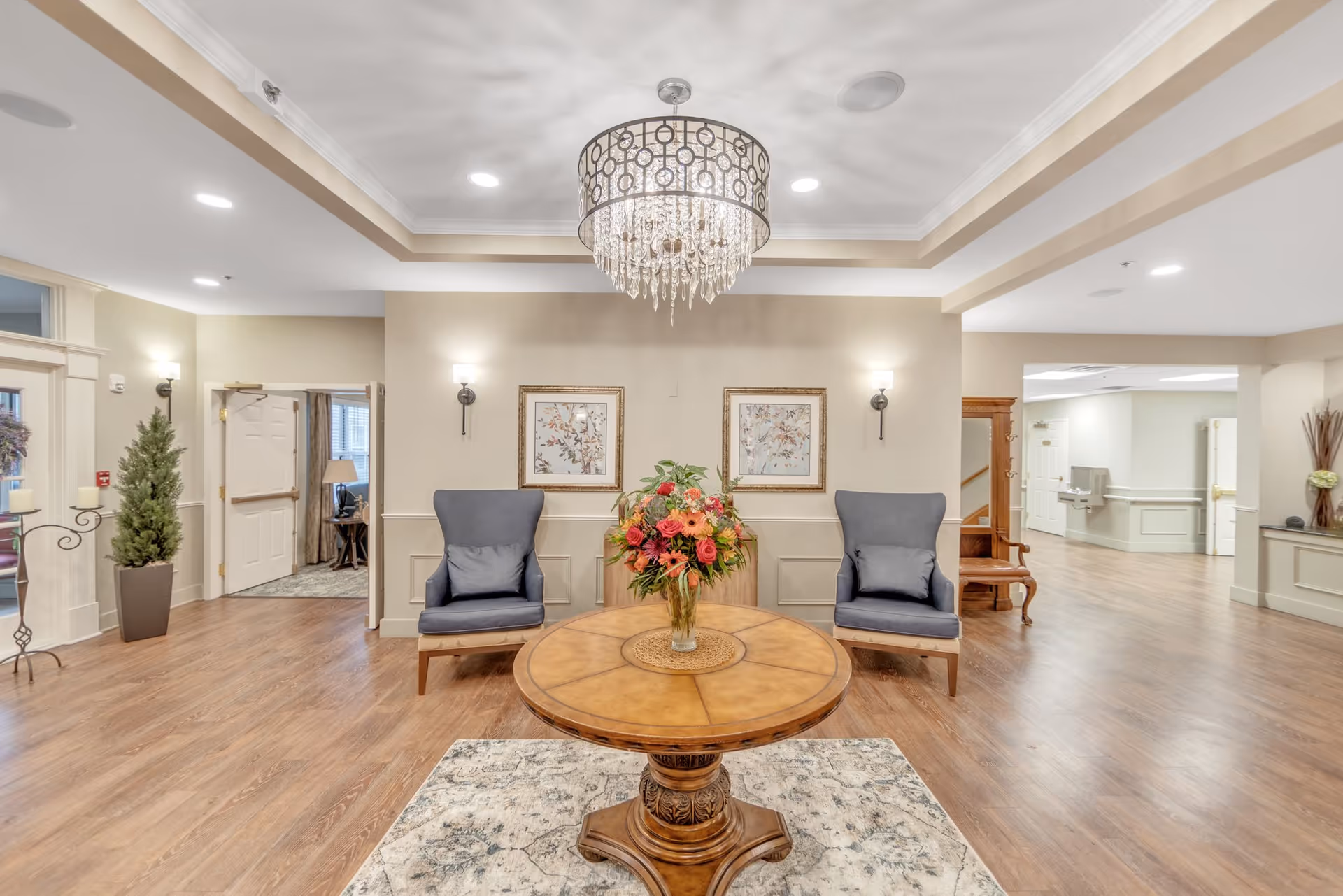 A spacious and well-lit interior common area with wooden flooring, a round wooden table with a floral arrangement in the center, two blue cushioned chairs against the wall, framed artwork above the chairs, and a decorative chandelier hanging from the ceiling. There are doorways leading to other rooms and hallways, with neutral-colored walls and soft lighting.