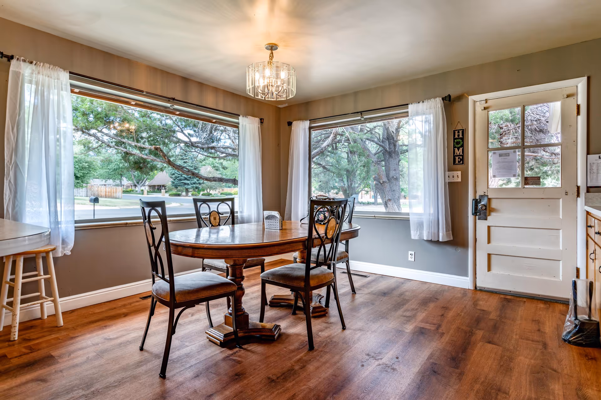 A dining area with a wooden table and four chairs with decorative backs. The room has large windows with white curtains, allowing a view of green trees outside. There is a white door with glass panels on the right side, and a small wooden stool is visible on the left. The floor is wooden, and a chandelier-style light fixture hangs from the ceiling.