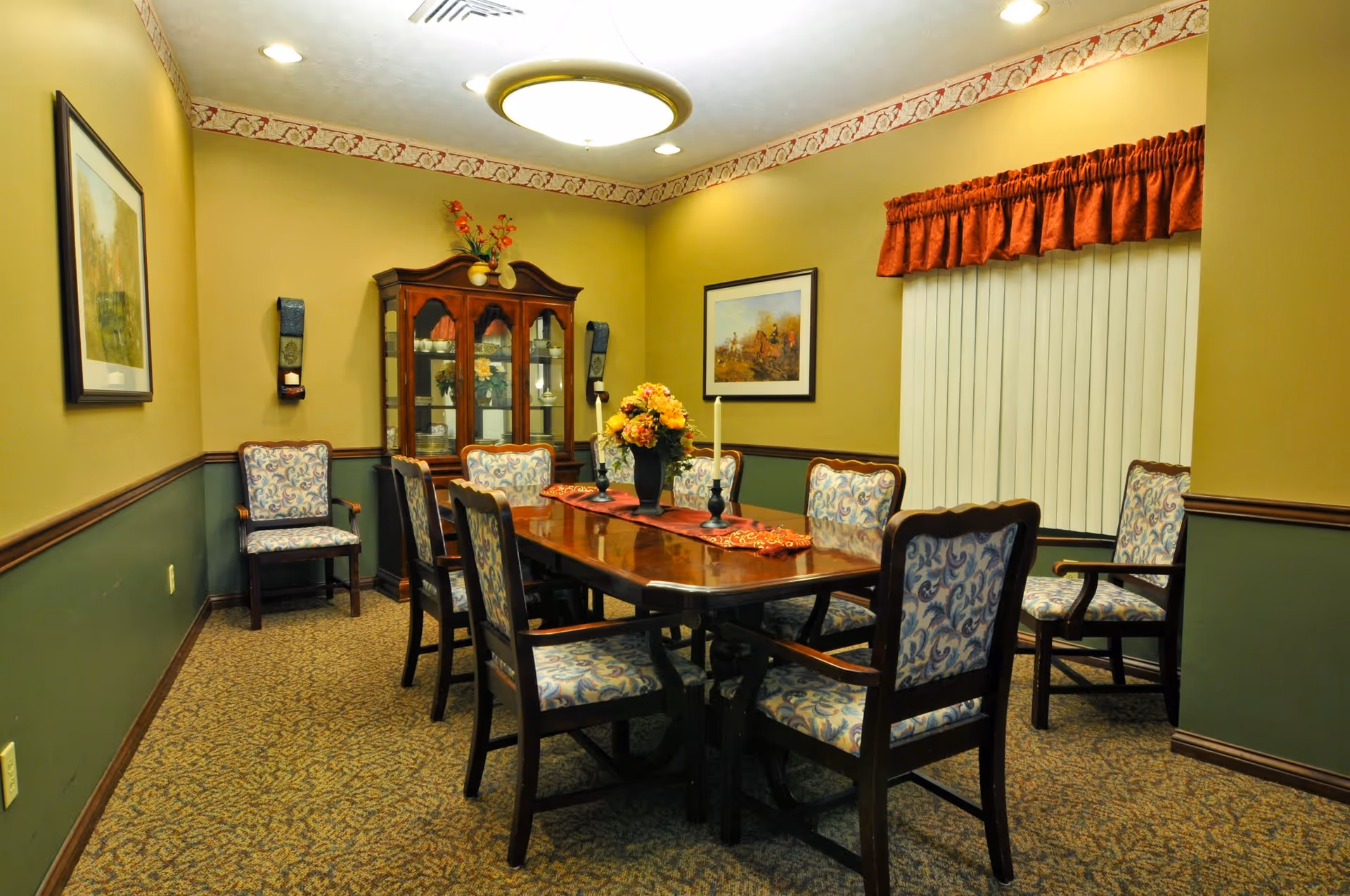 A dining room with a polished wooden table surrounded by upholstered chairs, a china cabinet, framed artwork and floral centerpieces.