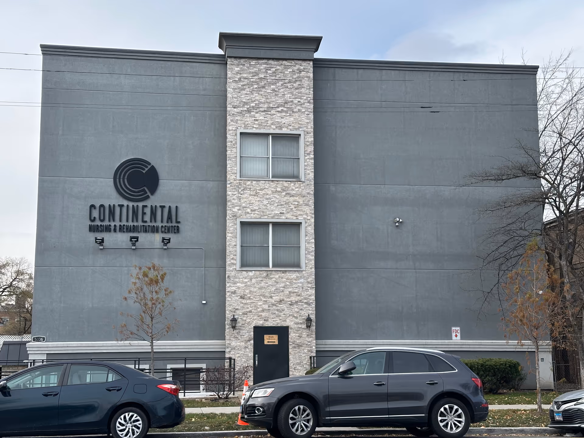 Exterior front view of the Continental Nursing & Rehabilitation Center building with a gray facade, stone accent in the middle, two windows, a black door labeled 'Main Entrance', and two parked cars in front on the street.
