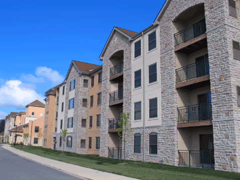 Exterior view of a multi-story residential building with stone facade and balconies along a street under a blue sky.