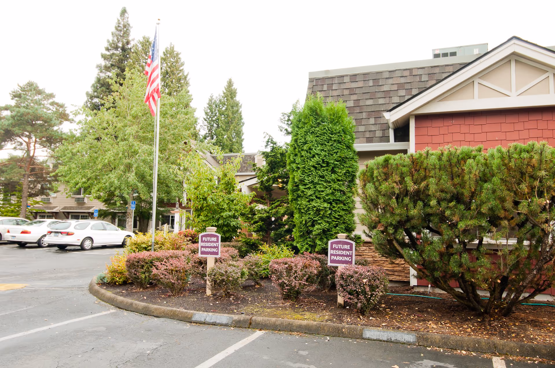 Parking area outside a building with landscaped bushes and trees, two signs indicating 'Future Resident Parking', an American flag on a flagpole, and several parked cars in the background.