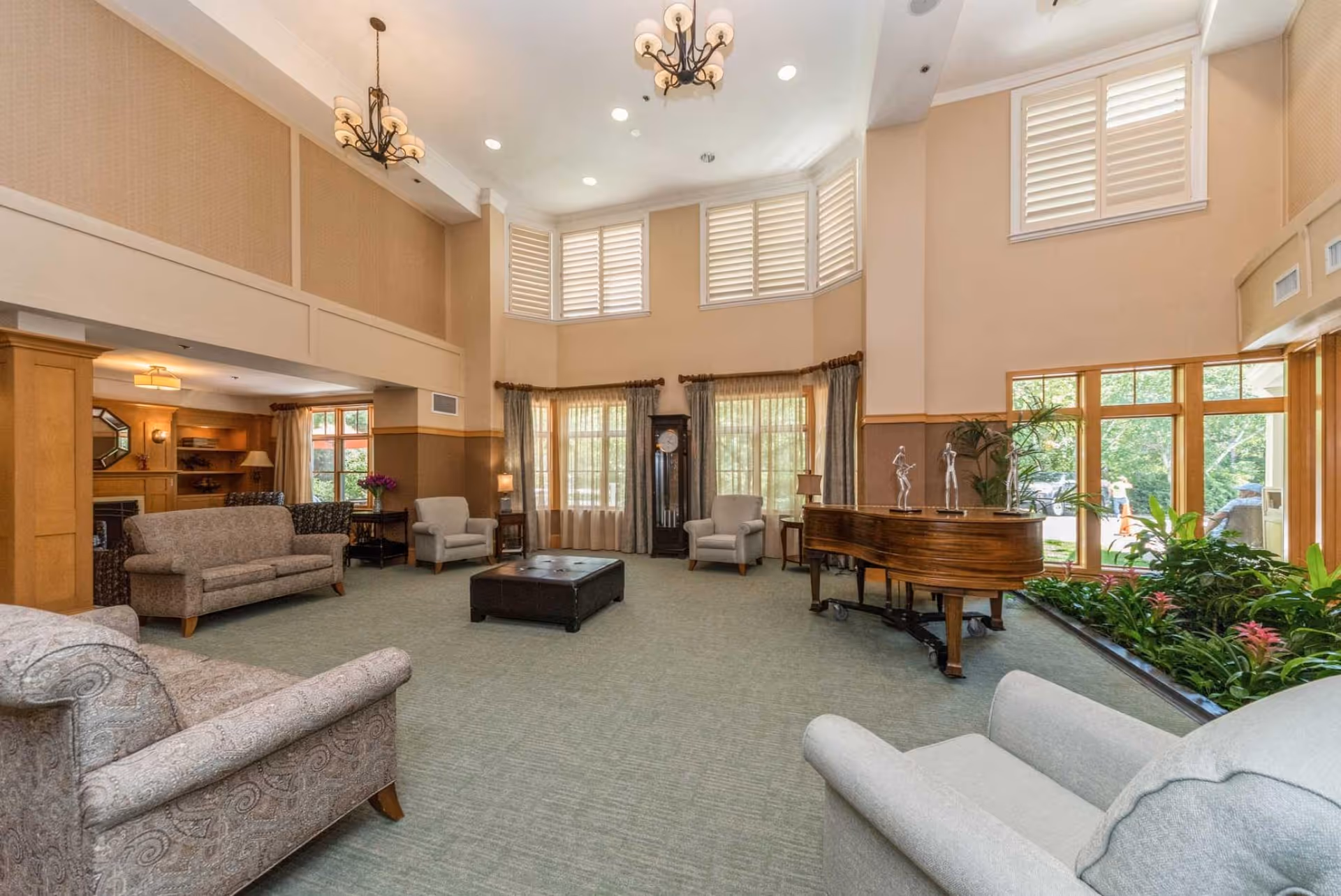 A spacious and well-lit living room area with high ceilings and large windows covered with white shutters. The room features multiple upholstered armchairs and sofas arranged around a central dark ottoman. There is a grand piano with decorative sculptures on top, a tall grandfather clock, and a variety of plants near the windows. The walls are painted beige with wooden paneling, and two chandeliers hang from the ceiling.