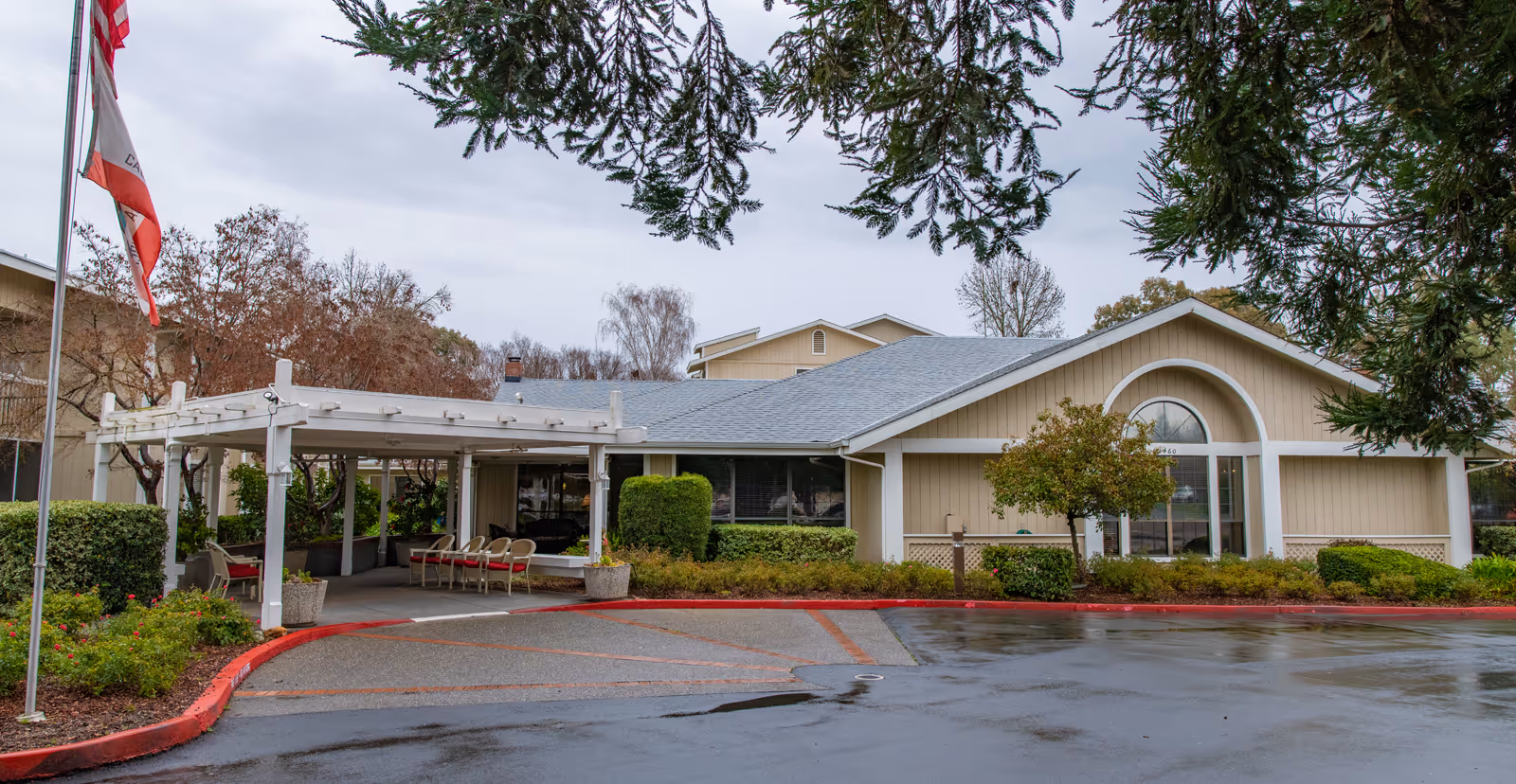 Front entrance of a single-story senior living building with a covered drop-off canopy, seating, landscaped shrubs and wet driveway under an overcast sky.