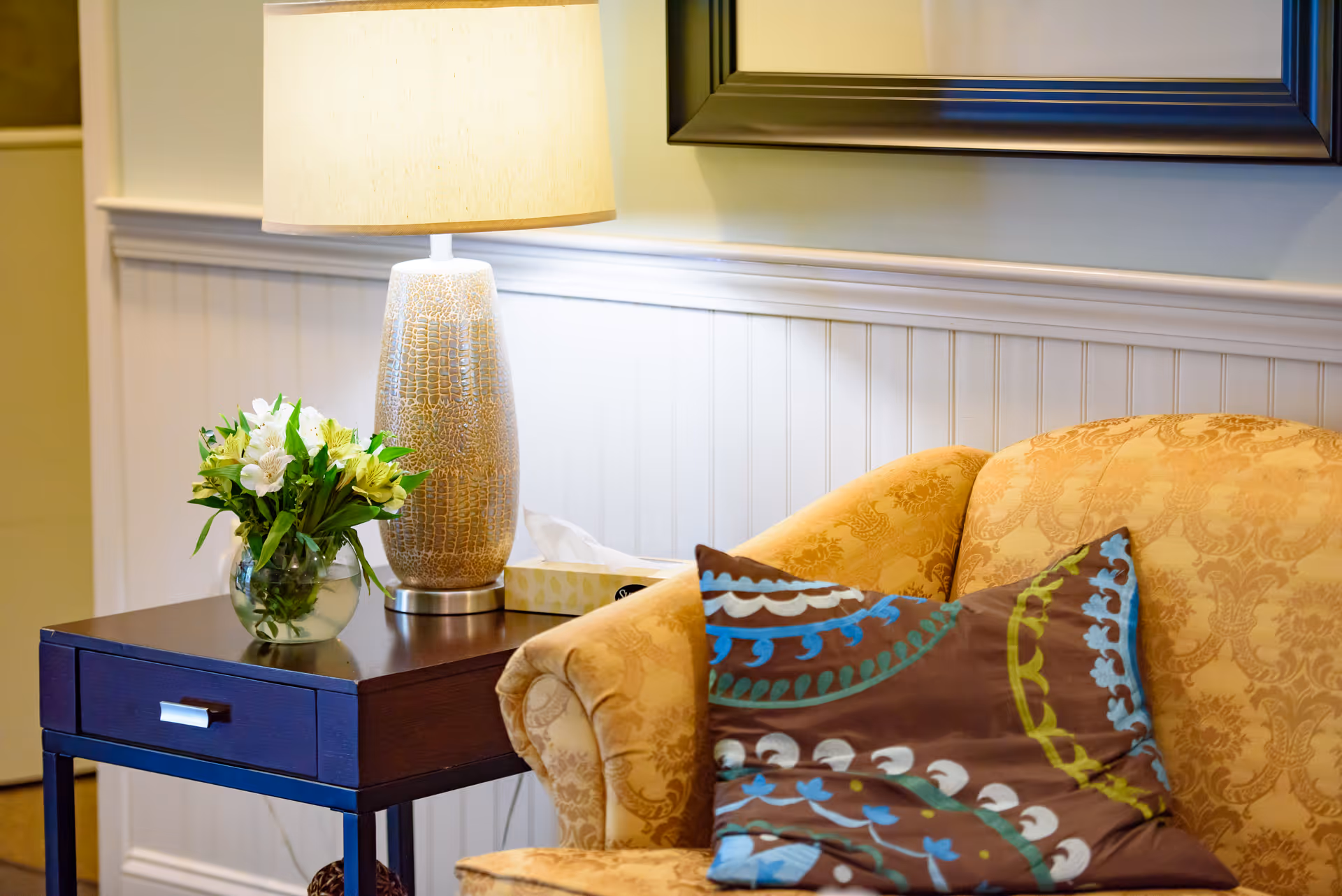 A cozy living room corner with a patterned beige armchair featuring a decorative pillow with blue, green, and white designs. Next to the chair is a dark wooden side table holding a textured lamp with a beige shade, a small glass vase with white and green flowers, and a box of tissues. The background shows white wainscoting on the wall and a framed picture partially visible above the chair.