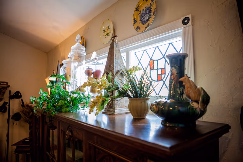 A wooden cabinet topped with decorative items including glass jars, a small potted plant, a tall triangular sculpture, and a ceramic vase with a bird figure. Behind the cabinet is a window with stained glass panels and decorative plates mounted on the wall above it. The room has textured beige walls and soft lighting.