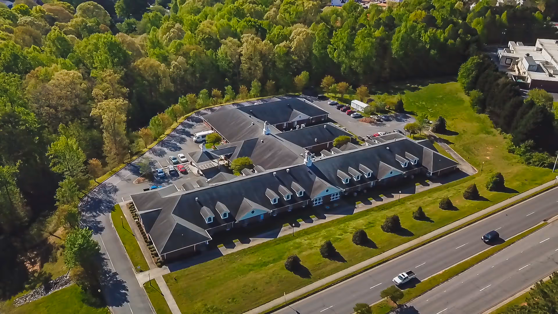 Aerial view of Terrace Ridge Assisted Living facility surrounded by trees and greenery, with a parking lot and a road with a few cars in front of the building.