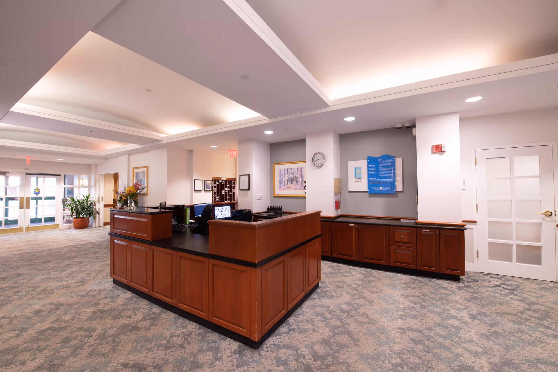 Reception area inside Youville House Assisted Living with a wooden front desk, carpeted floor, potted plants, framed artwork on the walls, and a clock. The space is well-lit with ceiling lights and has double glass doors leading outside.