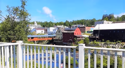 View of a small town with various buildings including a red covered bridge and a river flowing beneath it, seen from behind a white railing on a balcony or porch. Trees and greenery surround the area under a blue sky with a few clouds.