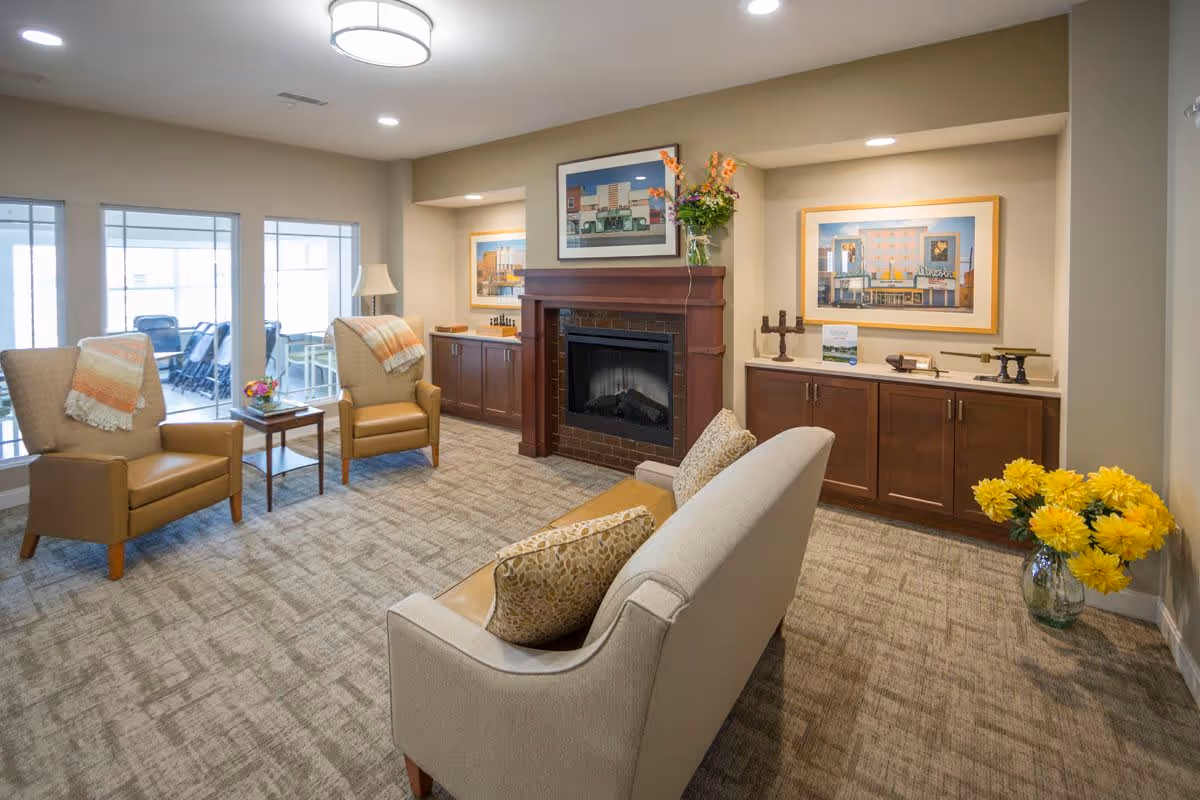 Comfortable senior living lounge with armchairs and a sofa arranged around a fireplace, cabinets, framed artwork, and a vase of yellow flowers.