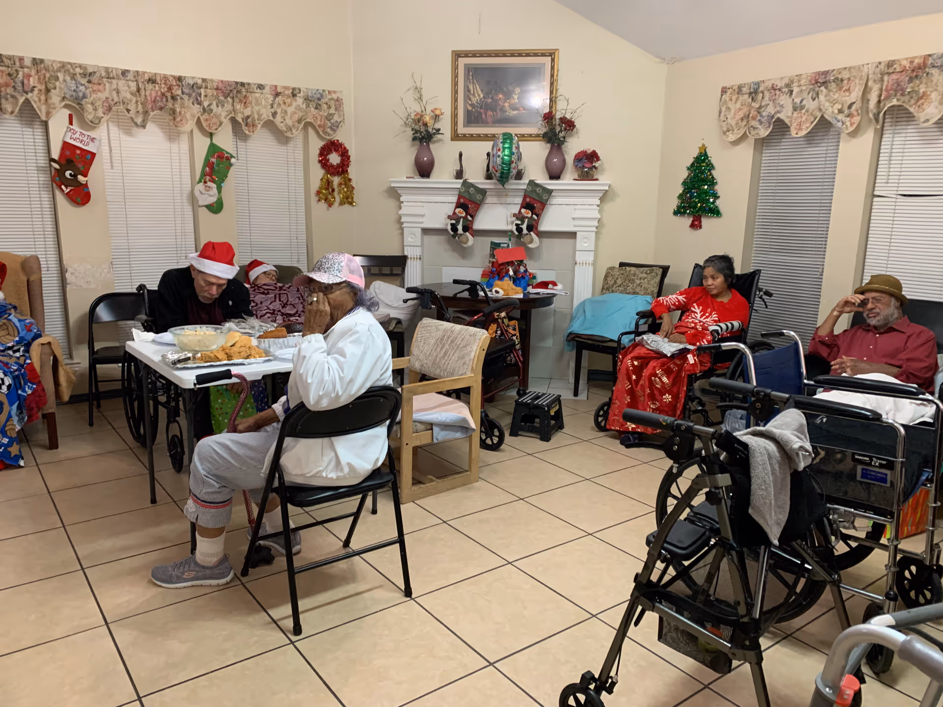A holiday-decorated communal living room in a senior care home with several elderly residents seated in chairs and wheelchairs around a table and near a fireplace.