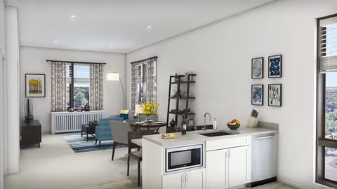 A bright and modern open-concept living space in a senior living facility featuring a small kitchen area with white cabinets, a microwave, and a sink. Adjacent to the kitchen is a dining table with chairs and a shelving unit. The living room area includes a blue sofa, a coffee table, and large windows with patterned curtains allowing natural light to fill the room. The walls are decorated with framed artwork.