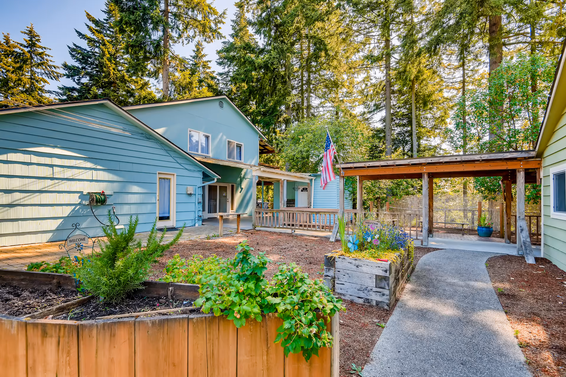 Outdoor view of a senior living facility courtyard with light blue buildings, a wooden pergola, garden beds with plants, an American flag, and tall trees in the background under a clear sky.