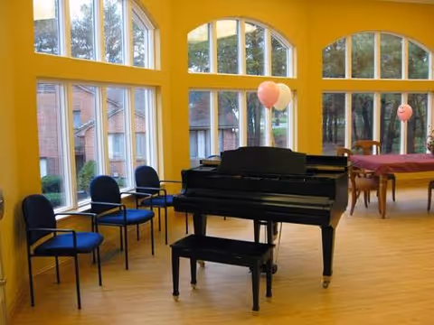 A bright room with large arched windows letting in natural light, featuring a black grand piano with a matching bench in the center. There are four blue chairs lined up against the wall on the left side and a wooden table with chairs and pink balloons in the background. The walls are painted yellow and the floor is wooden.