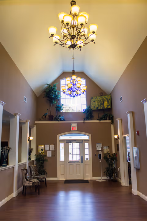 Interior view of a senior living facility entrance area with high vaulted ceilings, two large chandeliers, a window above the door, plants, chairs, and wall-mounted lights.