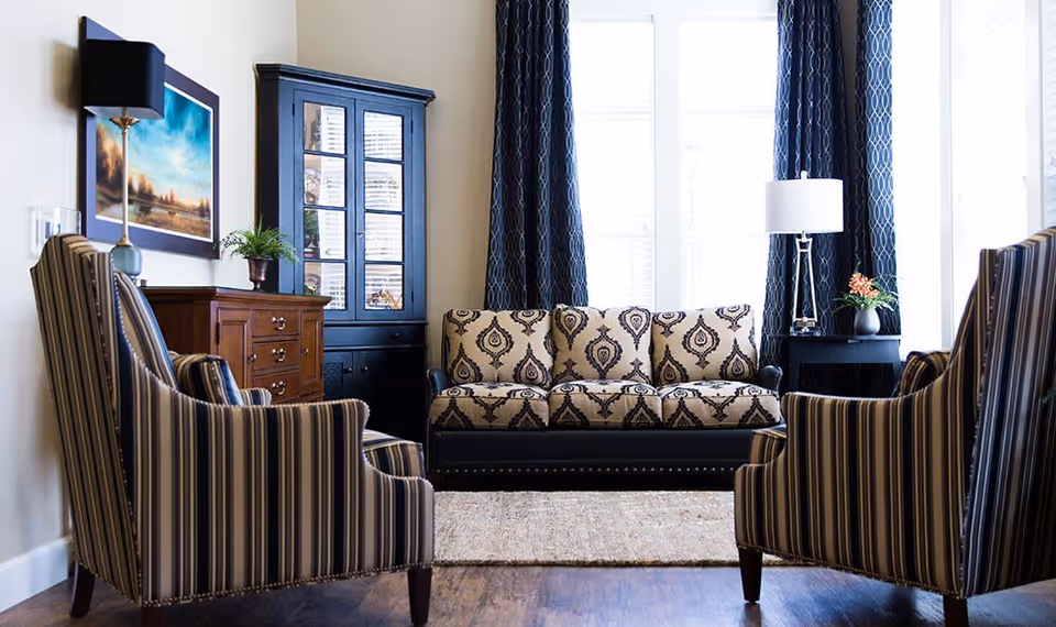 A well-lit living room with a patterned beige and black sofa centered against large windows with dark blue curtains. Two striped armchairs face the sofa, and there is a wooden cabinet with glass doors and a chest of drawers on the left side. A floor lamp with a black shade and a table lamp with a white shade are also visible, along with a small plant and a framed landscape painting on the wall.