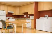 A kitchen area with wooden cabinets, a white refrigerator, stove, microwave, dishwasher, and a washing machine and dryer. There is a wooden table with a chair in the foreground. The walls are painted a warm reddish color.