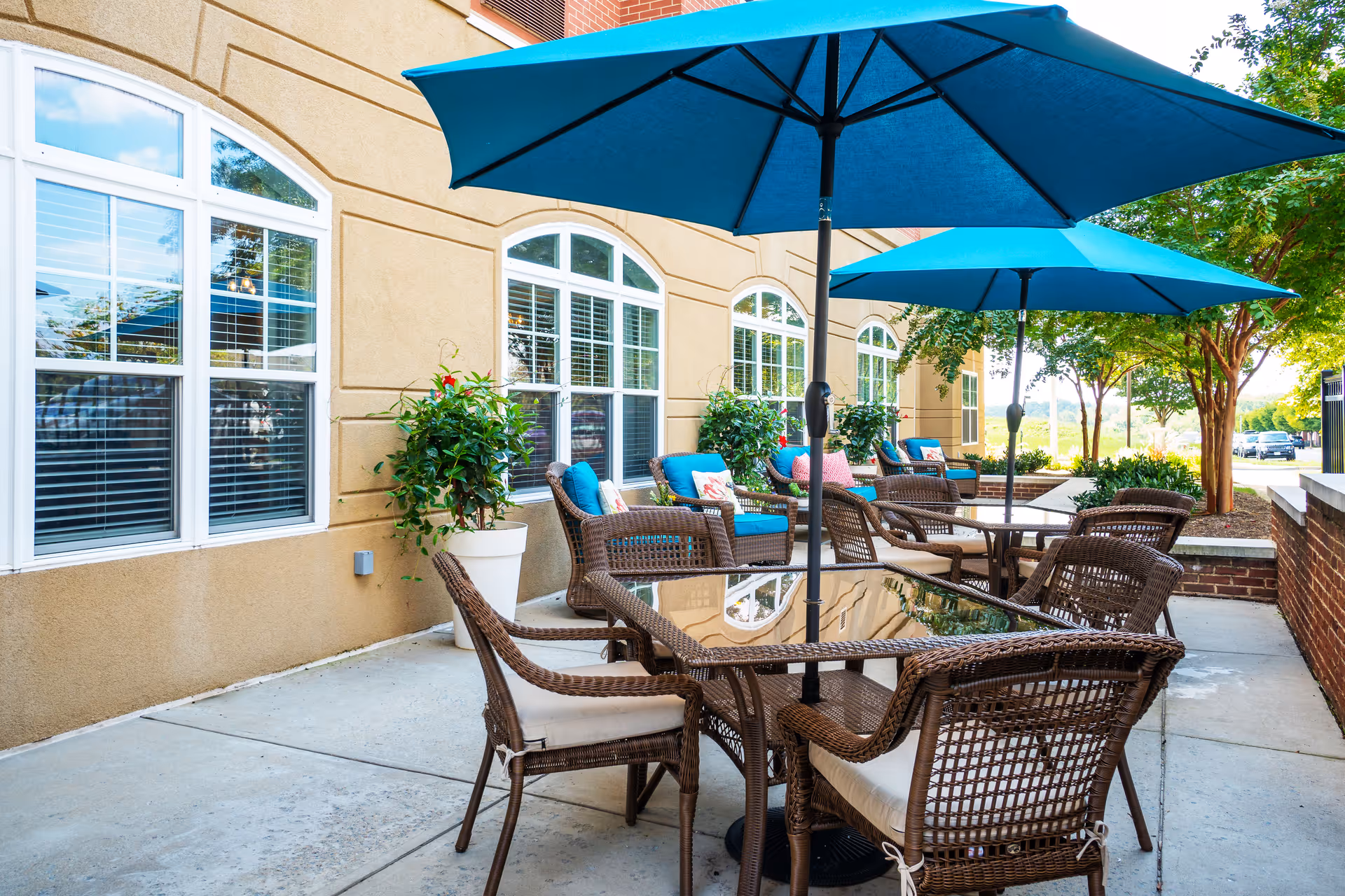 Outdoor patio area at RoseWood Village Assisted Living and Memory Care - Hollymead featuring wicker chairs with cushions around glass-top tables shaded by large blue umbrellas, potted plants, and a beige building with large windows in the background.