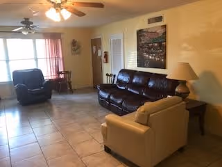 A living room with a brown leather sofa, a beige armchair, and a dark recliner chair. There is a ceiling fan with lights, a window with red curtains, a small wooden table with chairs near the window, a floor lamp beside the sofa, and a framed picture on the wall.