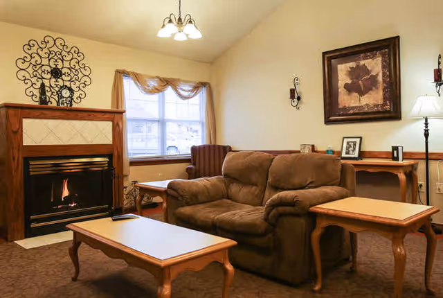 A cozy living room with a brown upholstered loveseat, a wooden coffee table, and side tables. There is a fireplace with a decorative metal wall piece above it, a window with sheer curtains, an armchair in the corner, and a framed floral artwork on the wall. A floor lamp and a ceiling light fixture provide lighting.