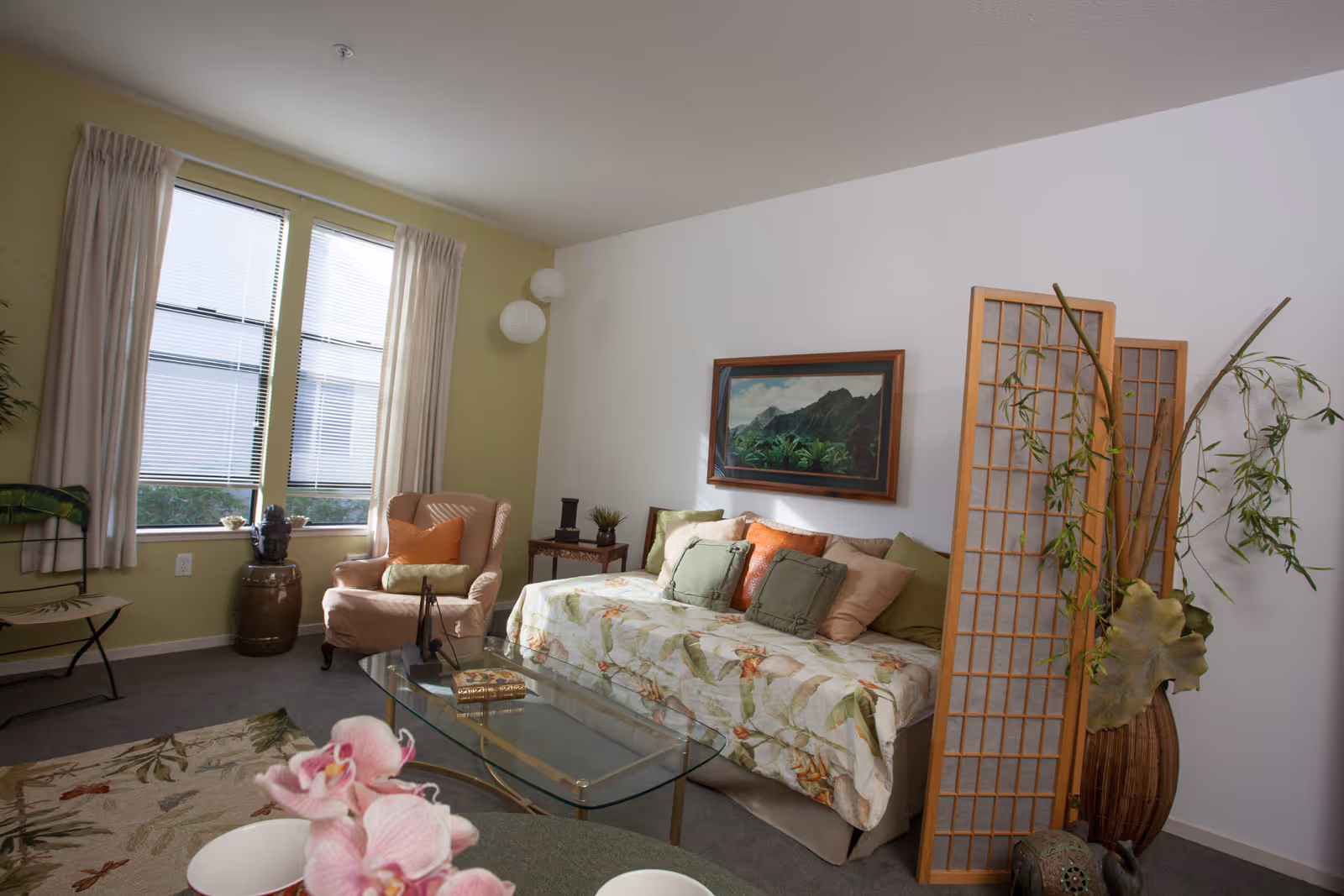 A cozy living room with a floral-patterned couch adorned with multiple pillows, a glass coffee table, an armchair with an orange pillow, a side table with decorative items, a large window with blinds and curtains, a framed landscape painting on the wall, and a wooden room divider with green plants in a vase beside it.