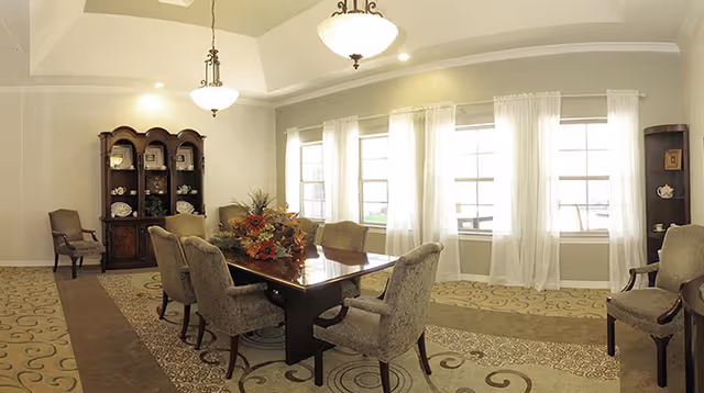 A well-lit dining room with a large wooden dining table surrounded by upholstered chairs. The room features a patterned carpet, two hanging light fixtures, large windows with sheer white curtains, and a wooden cabinet displaying decorative items.