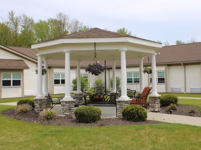 A white columned gazebo with chairs and hanging flower baskets in a grassy courtyard in front of a single-story care facility.