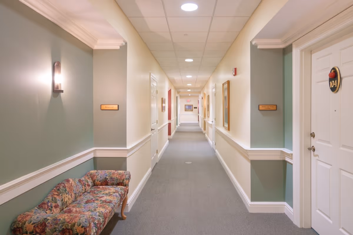 A long, well-lit hallway in a senior living facility with beige and green walls, white trim, and a gray carpet. There is a floral upholstered bench on the left side under a wall sconce light. Several doors and framed pictures line the hallway, including a door labeled 104 on the right.