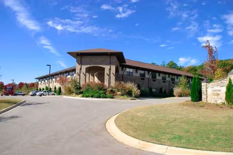 Two-story brick senior living facility with curved driveway and manicured landscaping under blue sky.