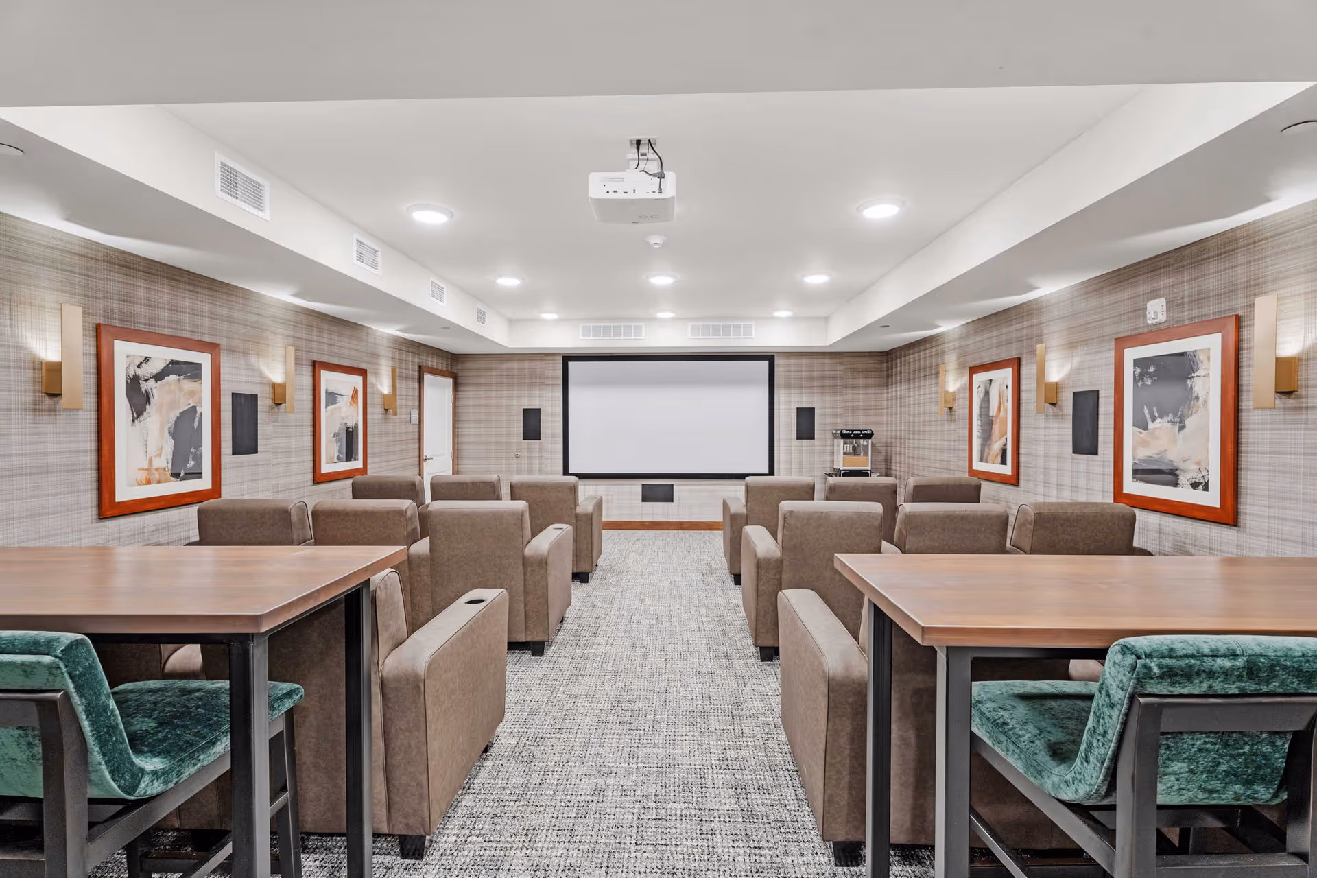 A modern media room with rows of brown armchairs facing a large projector screen on the far wall. The room has patterned wallpaper with framed abstract art on both side walls, recessed ceiling lights, and a projector mounted on the ceiling. There are two wooden tables with green upholstered chairs at the back of the room.