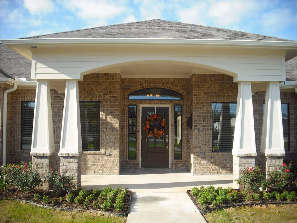 Front entrance of a brick building with a covered porch supported by white columns. The door has a decorative autumn wreath, and there are windows on either side of the door. Small bushes and plants line the walkway leading to the entrance.